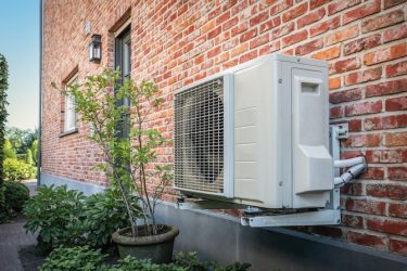 Air to air heat pump for cooling or heating the home. Side view of outdoor energy unit hanging on brick wall of beautiful house on a sunny day. Air conditioner or air heat pump. Outdoor unit powered by renewable energy.